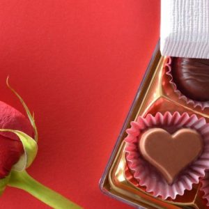 Gift detail of assorted chocolates and red rose on a red background with ribbon. Top view.
